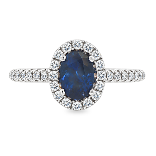 White gold ring with oval blue sapphire surrounded with a halo of diamonds on a diamond band. Shown on a clean white background.