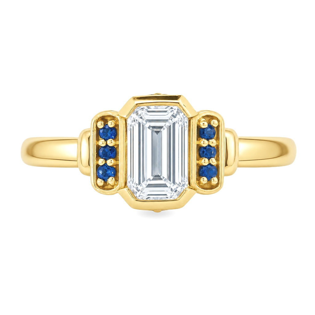 Yellow gold art deco ring with an emerald cut diamond an3 small blue sapphires on either side. Set on a white background.