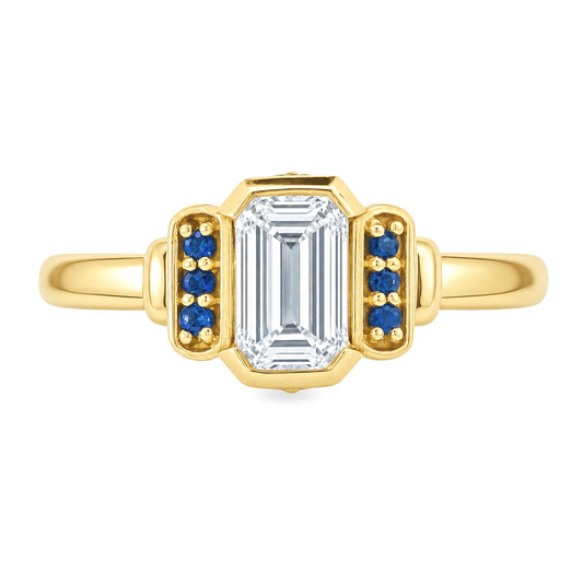 Yellow gold art deco ring with an emerald cut diamond an3 small blue sapphires on either side. Set on a white background.